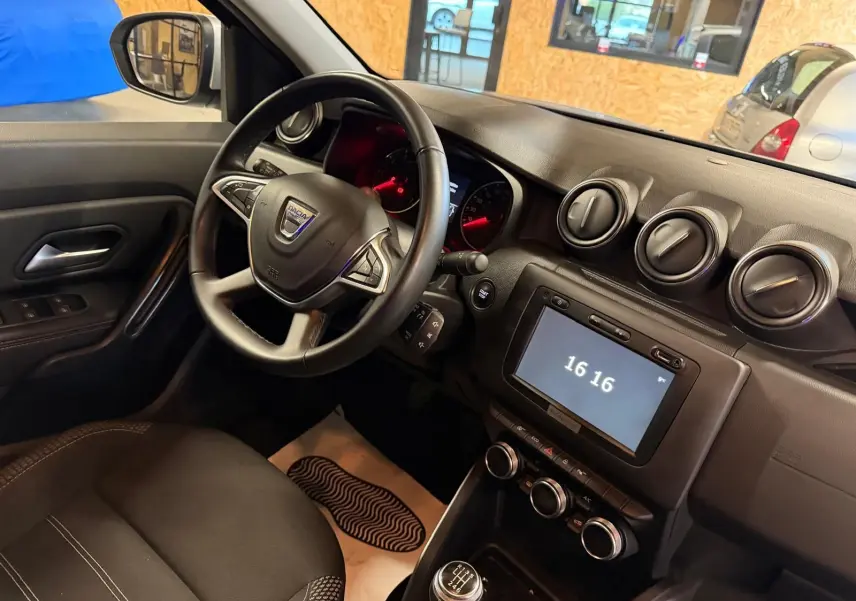 Vue intérieure côté conducteur du Dacia Duster gris 2019, volant multifonction et tableau de bord avec écran tactile allumé à 16h16.