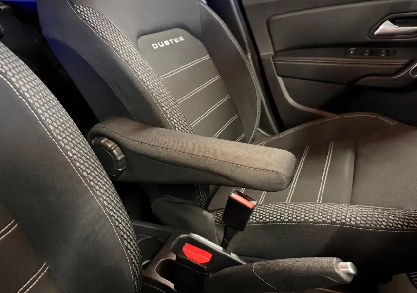 Gros plan sur le siège conducteur noir avec accoudoir et logo DUSTER, côté gauche intérieur d'un Dacia Duster gris.