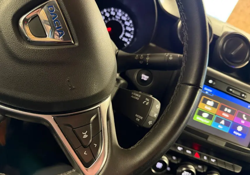 Gros plan sur le volant en cuir noir du Dacia Duster gris, avec commandes audio et tableau de bord lumineux en arrière-plan.
