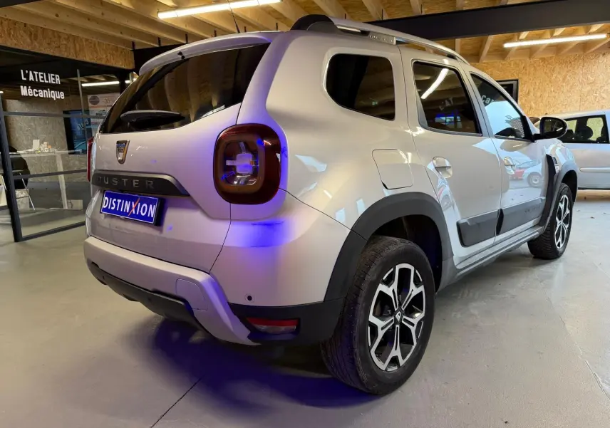 Vue 3/4 arrière droite du Dacia Duster gris 2019 avec jantes alliage et barres de toit en intérieur d'atelier.