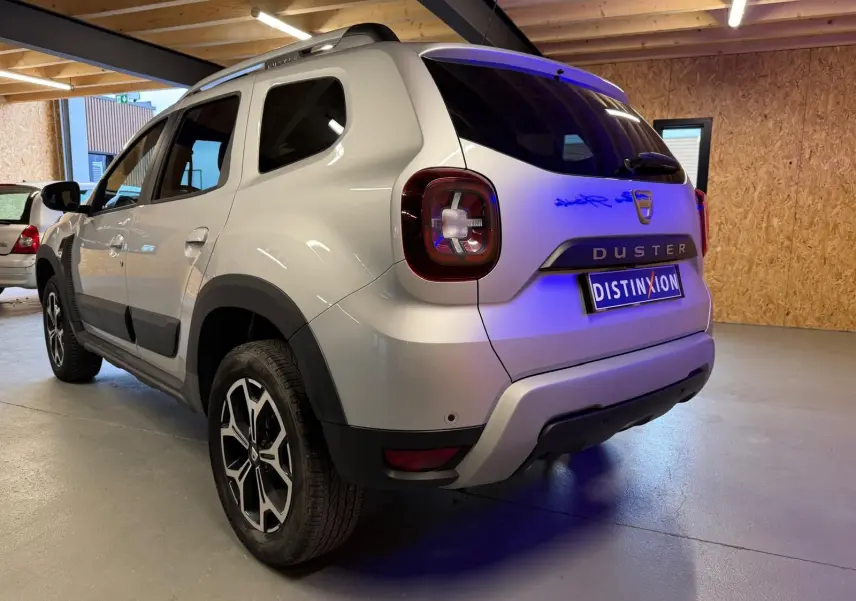 Vue 3/4 arrière droit d'un Dacia Duster gris 2019 avec jantes alliage et barres de toit en intérieur d'atelier.