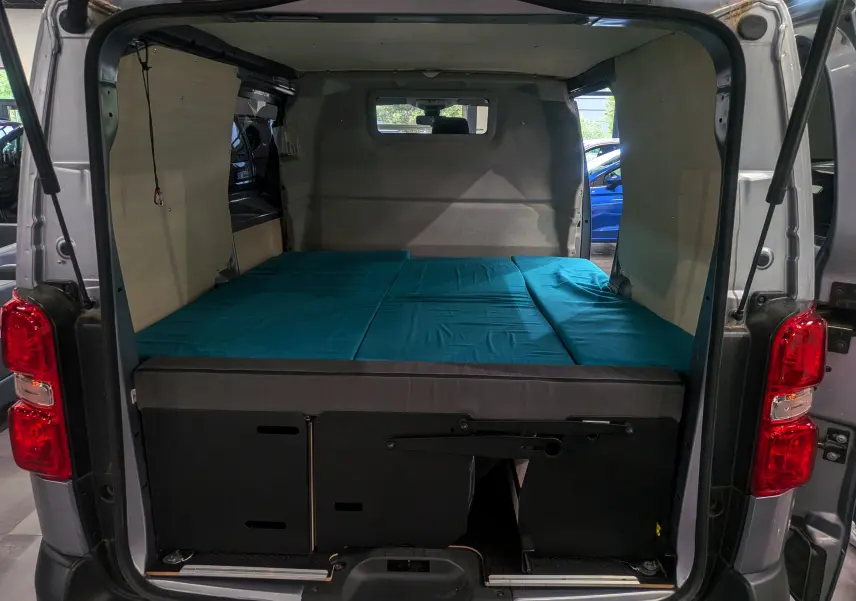 Vue arrière ouverte du Peugeot Expert Fourgon gris acier 2023, aménagé avec un lit bleu et habillage bois intérieur.