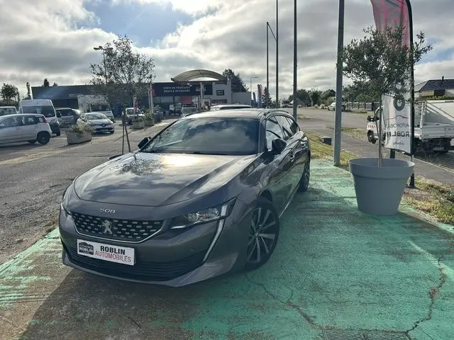 Peugeot 508 SW gris vue 3/4 avant droit, stationnée en extérieur avec calandre distinctive et jantes noires.