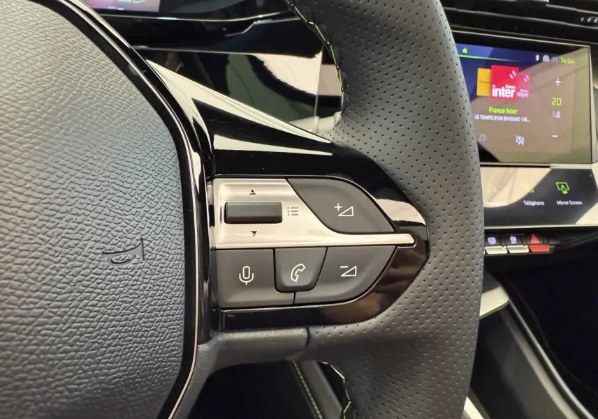 Gros plan sur le volant cuir perforé de la Peugeot 308 Hybrid gris foncé, avec commandes vocales et écran tactile en arrière-plan.