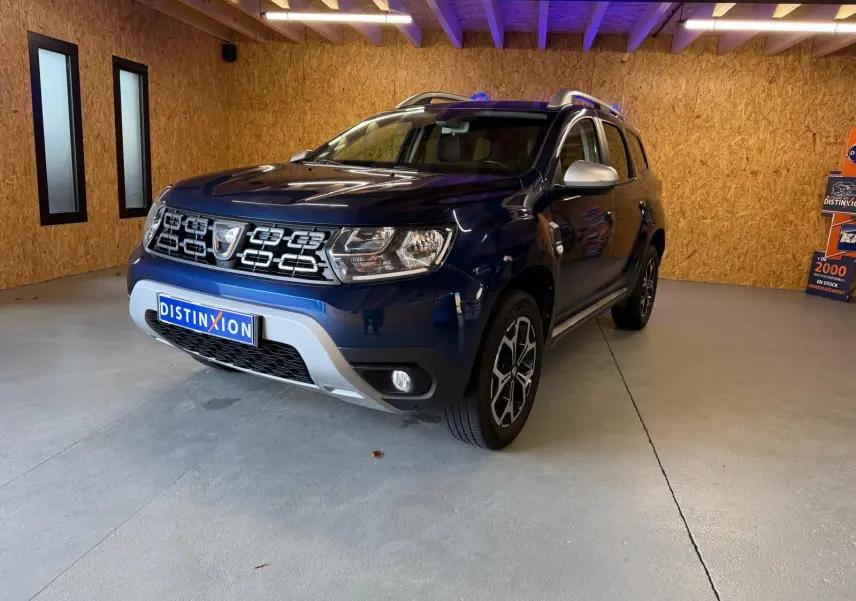 Vue 3/4 avant d'un Dacia Duster bleu avec calandre chromée et jantes alliage 17 pouces dans un garage.