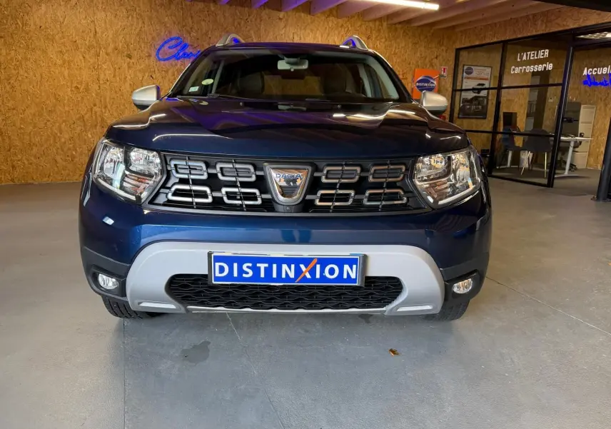Vue frontale d'un Dacia Duster bleu avec calandre noire et éléments chromés, stationné en intérieur showroom.