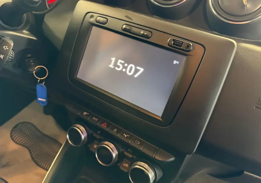 Vue rapprochée de la console centrale du Dacia Duster bleu, avec écran tactile affichant l'heure et commandes climatisation.