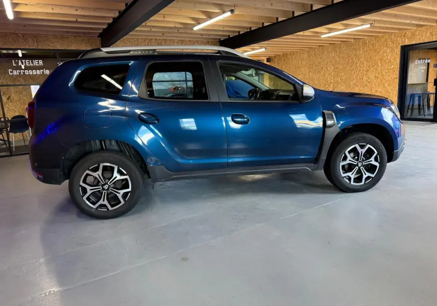 Vue latérale côté gauche d'un Dacia Duster bleu avec jantes alliage 17 pouces dans un atelier carrosserie.