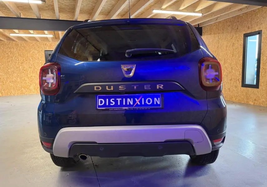 Vue arrière d'un Dacia Duster bleu avec logo distinctif et plaque d'immatriculation "DISTINXION" en intérieur lumineux.