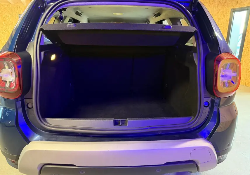 Coffre ouvert vu de l'arrière d'un Dacia Duster bleu, avec hayon et feux arrière visibles.