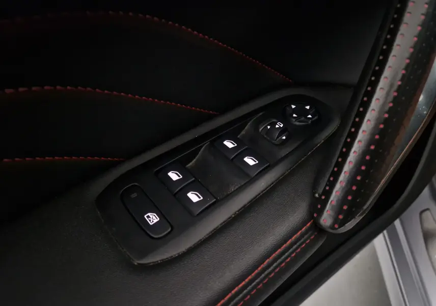 Gros plan sur les commandes de lève-vitres et rétroviseurs côté conducteur, cuir noir avec surpiqûres rouges.