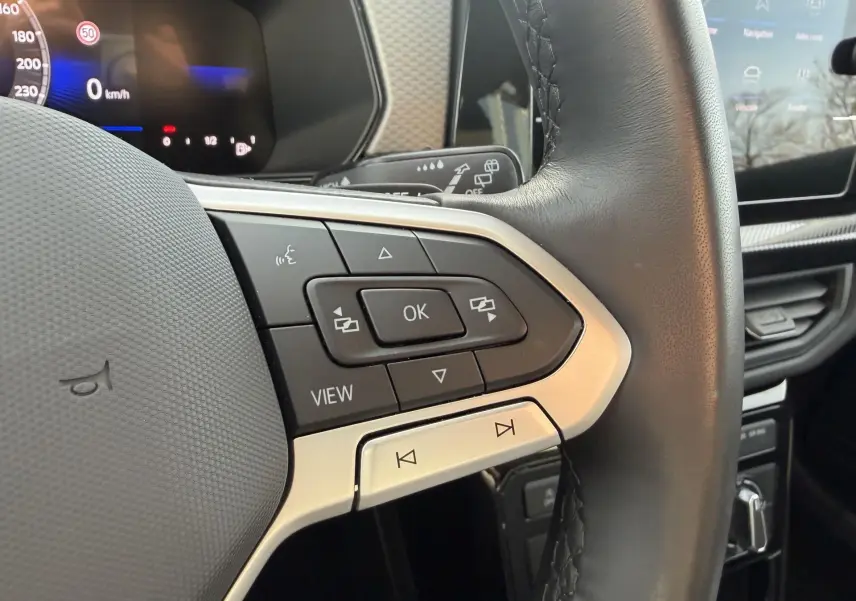 Gros plan sur les commandes multifonctions du volant cuir noir du Volkswagen T-Cross gris, avec écran digital en arrière-plan.