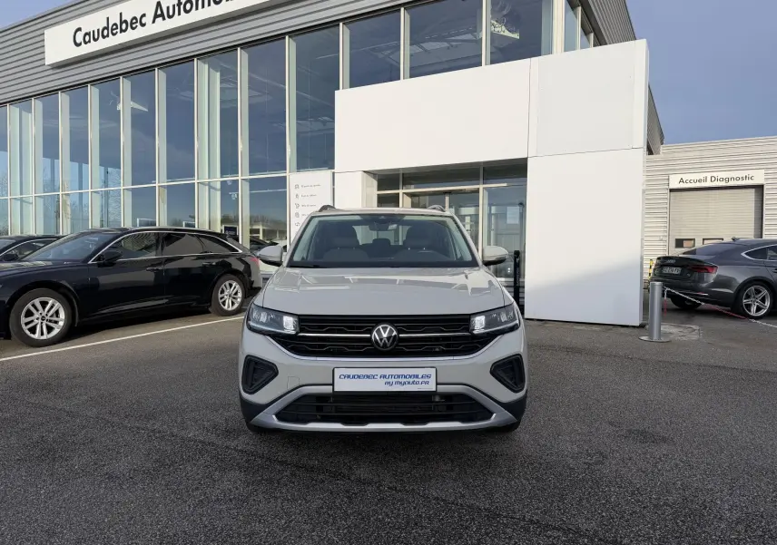 Vue avant d'un Volkswagen T-Cross gris garé devant un bâtiment de concession automobile par temps clair.