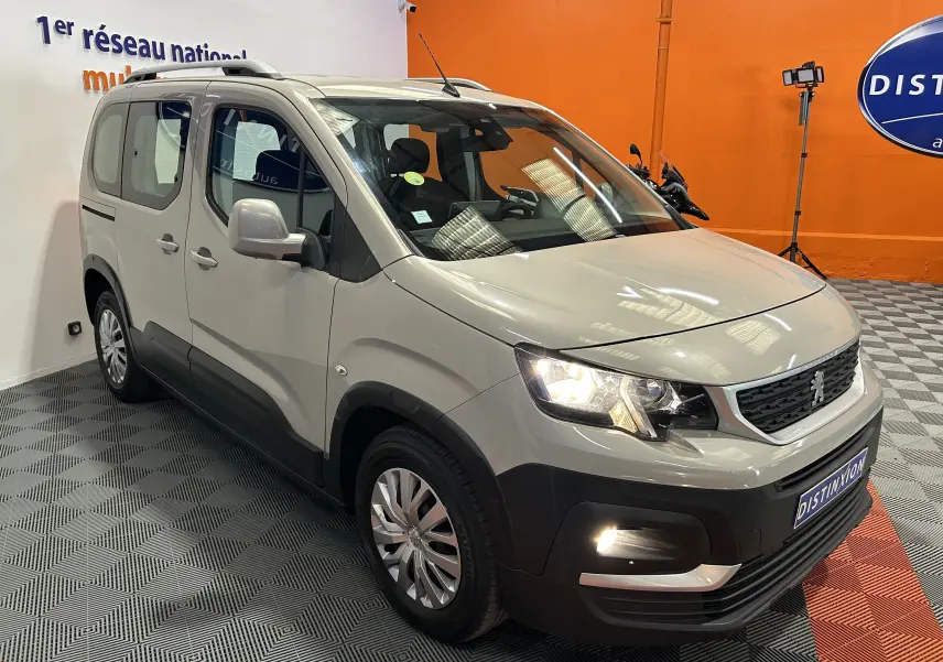 Peugeot Rifter gris sable en 3/4 avant droit, avec phares allumés et attelage visible dans un showroom orange.