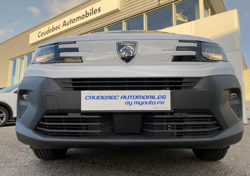 Vue frontale basse du Peugeot Partner Fourgon blanc avec logo lion et calandre noire, devant un bâtiment commercial.