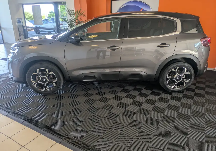 Profil latéral gauche d'un Citroën C5 Aircross hybride 2024 gris platinium avec toit noir, exposé en intérieur sur sol damier.