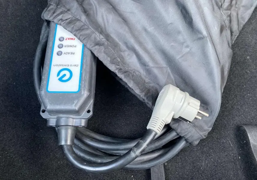 Gros plan sur le câble de recharge électrique noir avec boîtier blanc marqué "Zero Emission" dans le coffre d'une Nissan Leaf blanche.