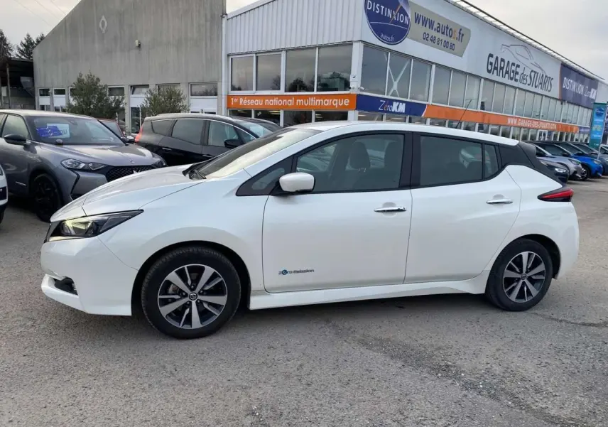 Profil côté gauche d'une Nissan Leaf 40 N-CONNECTA blanche, électrique, stationnée devant un garage multimarque.