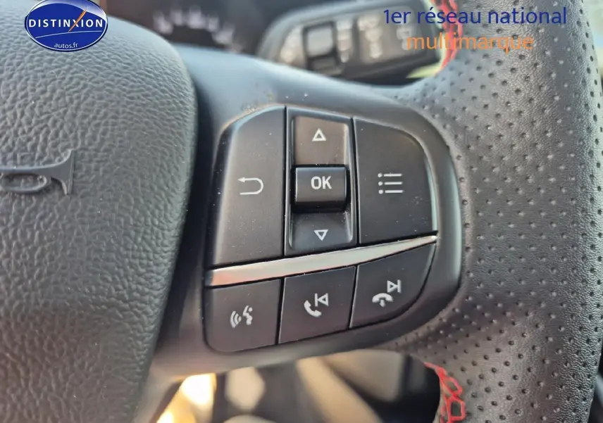 Gros plan sur les commandes multifonctions du volant cuir perforé avec surpiqûres rouges d'une Ford Focus gris magnétique.