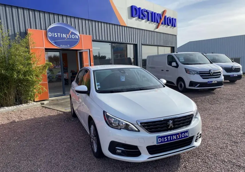 Peugeot 308 blanc nacré vue 3/4 avant droit devant un concessionnaire Distinxion par temps clair.