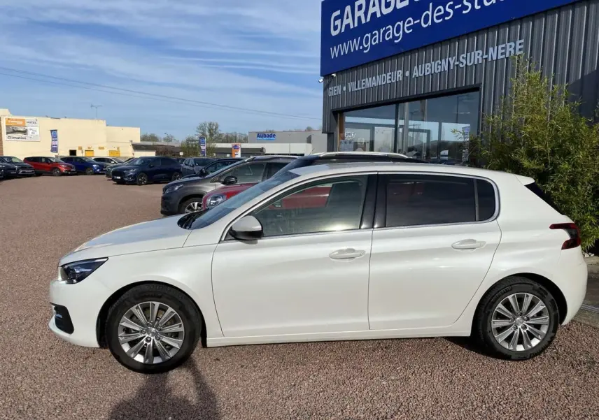 Profil côté gauche d'une Peugeot 308 blanche nacrée 2017 garée devant un garage avec vitres surteintées.