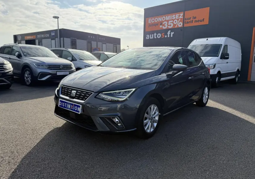SEAT Ibiza 1.0 EcoTSI 95 Xcellence gris Magnetic Tech vue 3/4 avant sur parking avec panneau promotionnel en arrière-plan