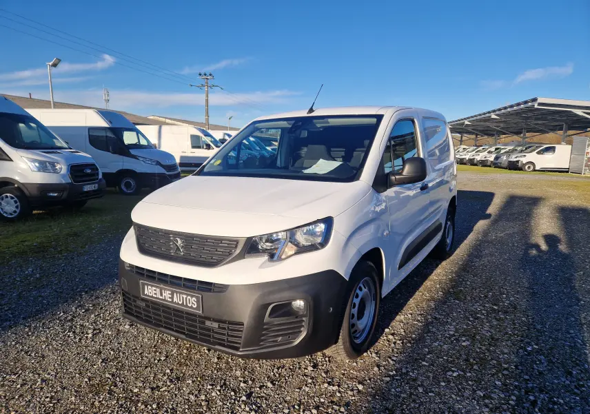 Vue 3/4 avant droite d’un Peugeot Partner blanc 2021 avec pare-chocs noir et calandre Peugeot, stationné sur un sol gravillonné.