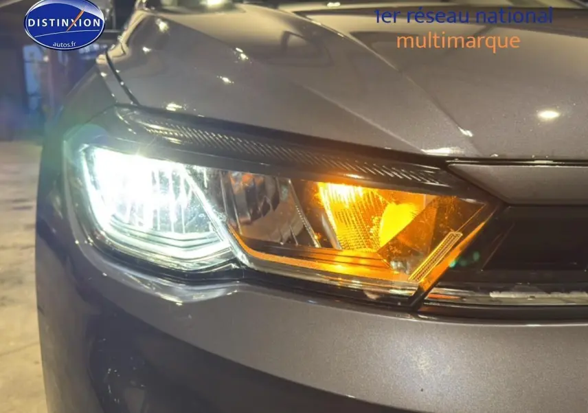 Gros plan sur le phare avant gauche allumé d'une Volkswagen Polo gris cendre, avec clignotant orange visible.