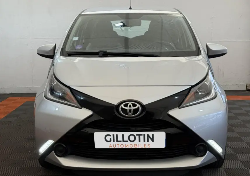 Vue avant frontale d'une Toyota Aygo gris clair avec calandre noire en forme de X et feux de jour LED allumés.