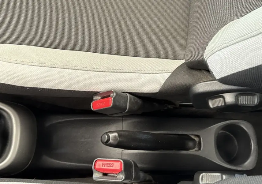 Vue rapprochée de la console centrale d'une Toyota Aygo gris clair, montrant le frein à main et les boucles de ceinture rouge.