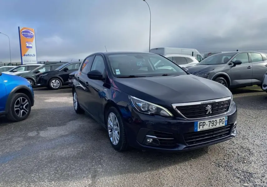 Peugeot 308 bleu foncé vue 3/4 avant droit sur parking avec calandre chromée et feux LED allumés.