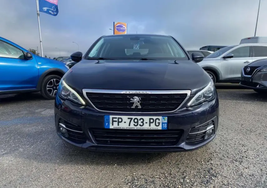 Peugeot 308 bleu foncé vue de face, avec calandre chromée et feux LED allumés, stationnée en extérieur.