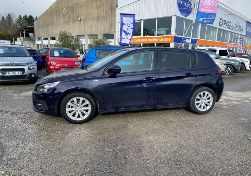 Peugeot 308 bleu foncé vue de profil côté gauche, garée devant un garage multimarque en extérieur.