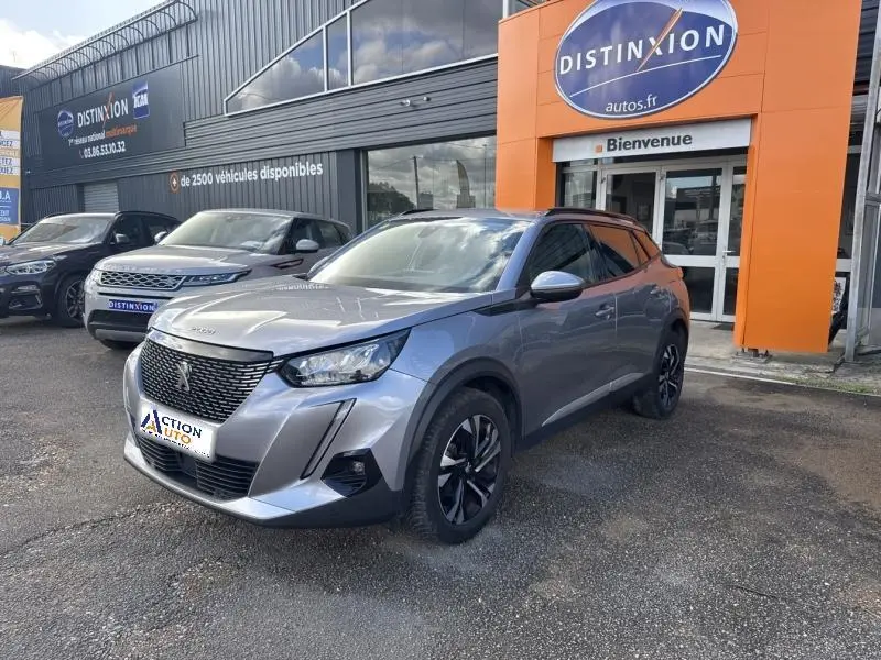 Peugeot 2008 gris Artense vue 3/4 avant droit, avec jantes alliage et calandre noire, stationnée devant un concessionnaire.