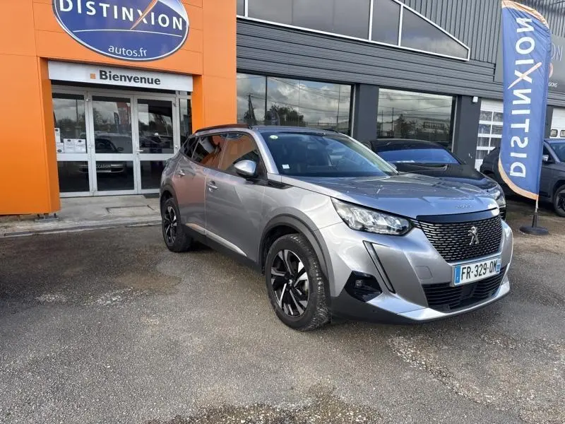 Peugeot 2008 gris Artense vue 3/4 avant droit, avec jantes alliage noires et calandre noire, devant un bâtiment orange.