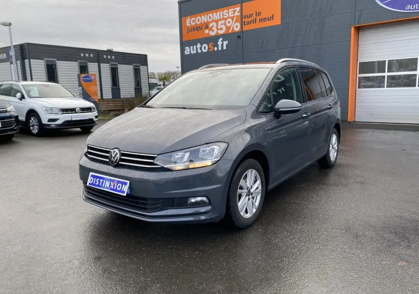Volkswagen Touran gris dauphin en 3/4 avant droit sur parking devant un bâtiment commercial.