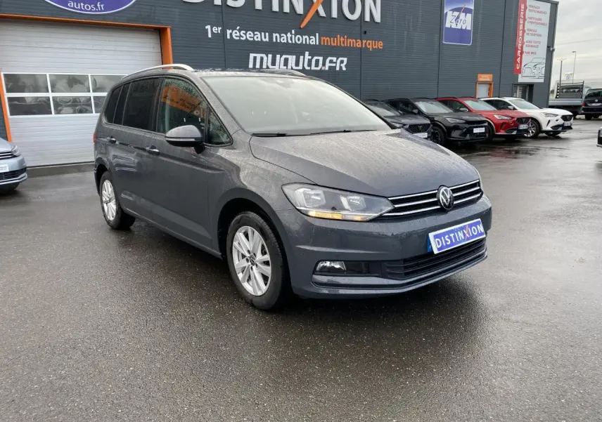 Volkswagen Touran gris dauphin vue 3/4 avant droit sur parking devant un bâtiment commercial.