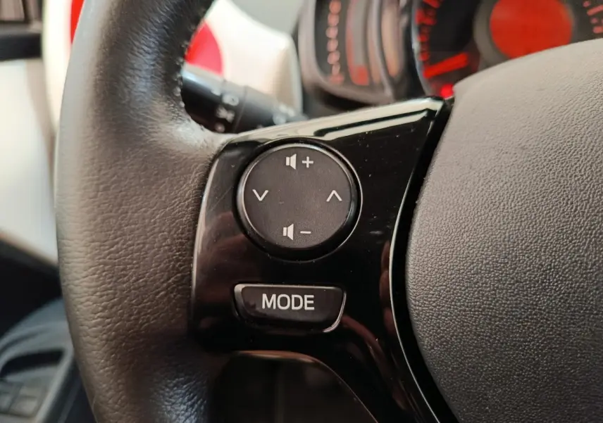Gros plan sur les commandes audio au volant cuir noir de la Toyota Aygo 2016, avec tableau de bord rouge en arrière-plan.