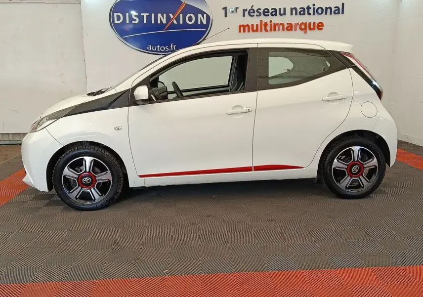 Profil côté gauche d'une Toyota Aygo blanche 2016 avec jantes noires et détails rouges, en intérieur showroom.