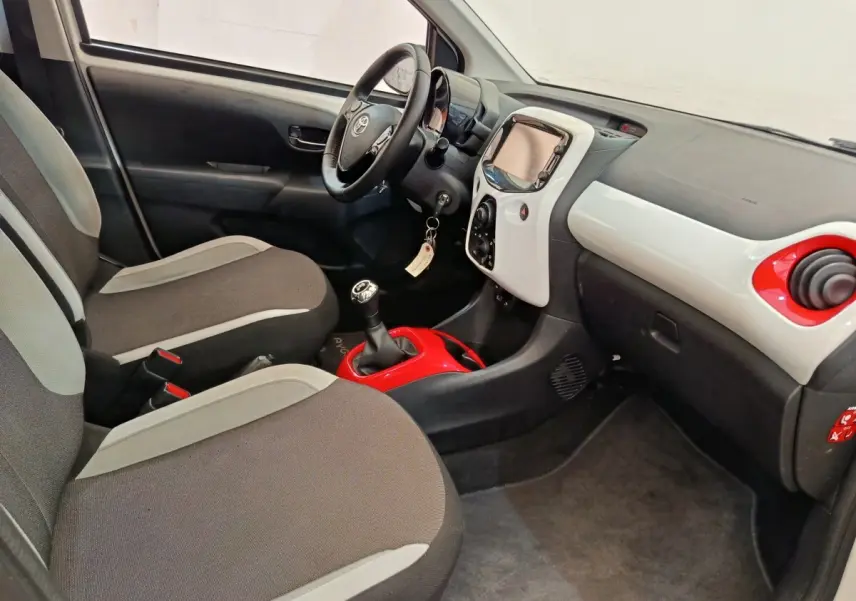Vue intérieure côté conducteur de la Toyota Aygo blanche, avec tableau de bord bicolore et levier de vitesses manuel rouge.