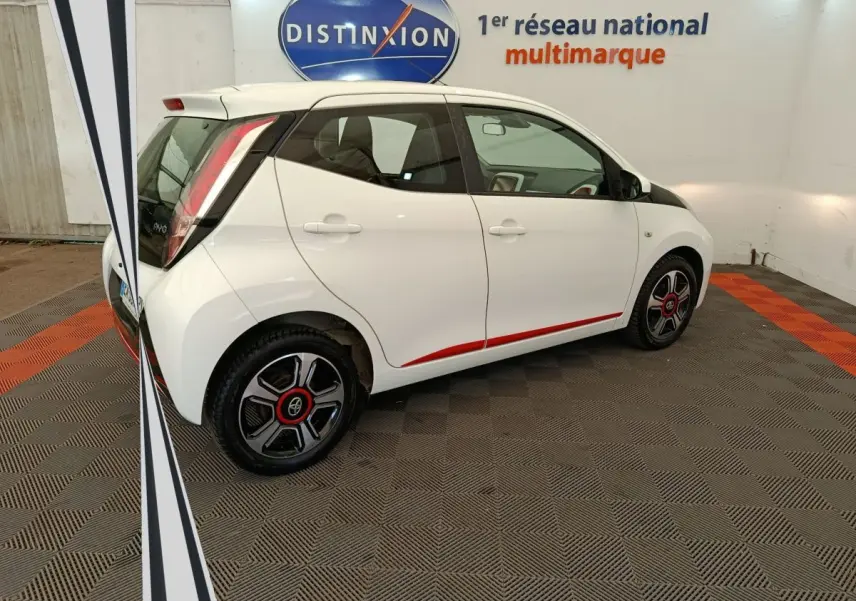 Vue 3/4 arrière droite d'une Toyota Aygo blanche avec jantes noires et rouges, soulignée d'une bande rouge sur le bas de caisse.