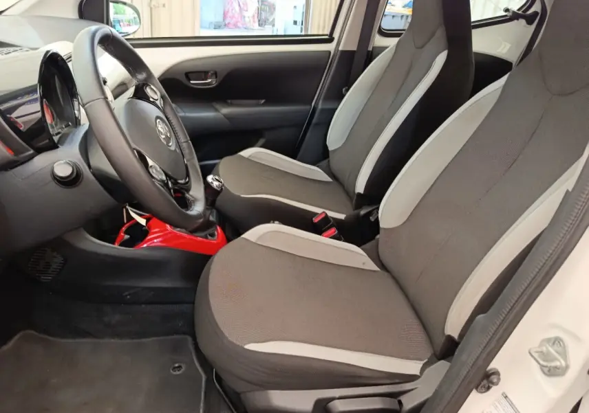 Vue intérieure côté conducteur de la Toyota Aygo blanche 2016, sièges gris avec console centrale rouge visible.