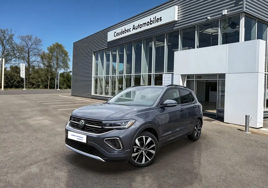 Volkswagen T-Cross gris en 3/4 avant droit, avec jantes alliage noires et grises, devant un bâtiment de concession.