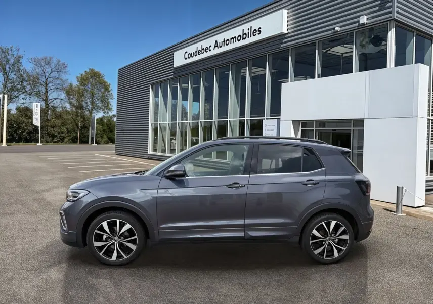 Vue de profil côté gauche d'un Volkswagen T-Cross gris 2025 avec jantes alliage 18 pouces devant un bâtiment commercial.