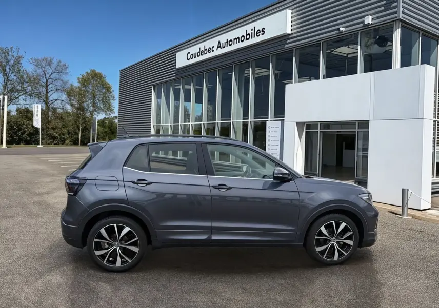 Vue latérale droite d'un Volkswagen T-Cross gris 2025 avec jantes alliage noires et détails chromés, devant un concessionnaire.