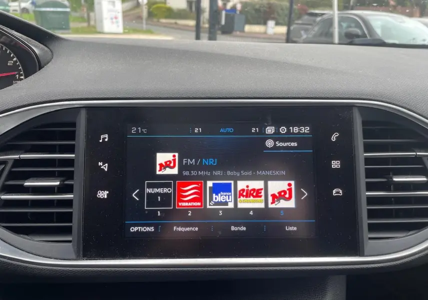 Écran tactile central de la Peugeot 308 gris Hurricane affichant la radio FM NRJ avec commandes tactiles et aérateurs latéraux.