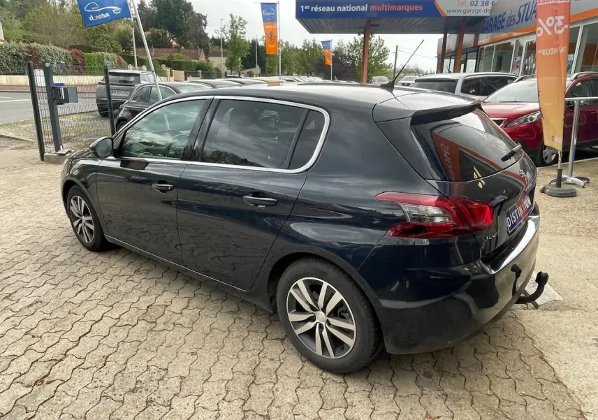 Vue 3/4 arrière droite d'une Peugeot 308 gris hurricane avec attelage de remorque, stationnée devant un garage.