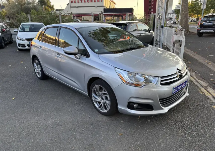 Citroën C4 gris clair vue 3/4 avant droit, stationnée devant un restaurant avec reflets métallisés.