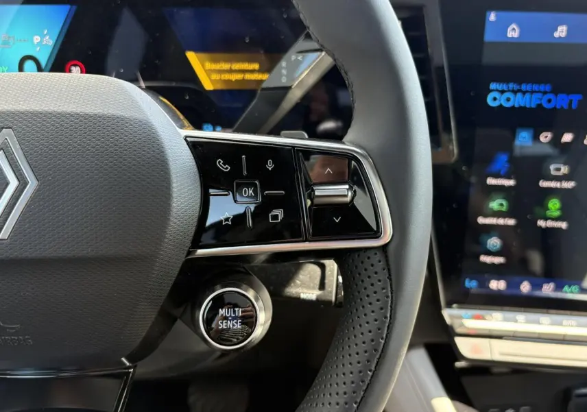 Gros plan sur le volant noir perforé et les commandes multifonctions du Renault Espace VI hybride, écran multifonction en arrière-plan.