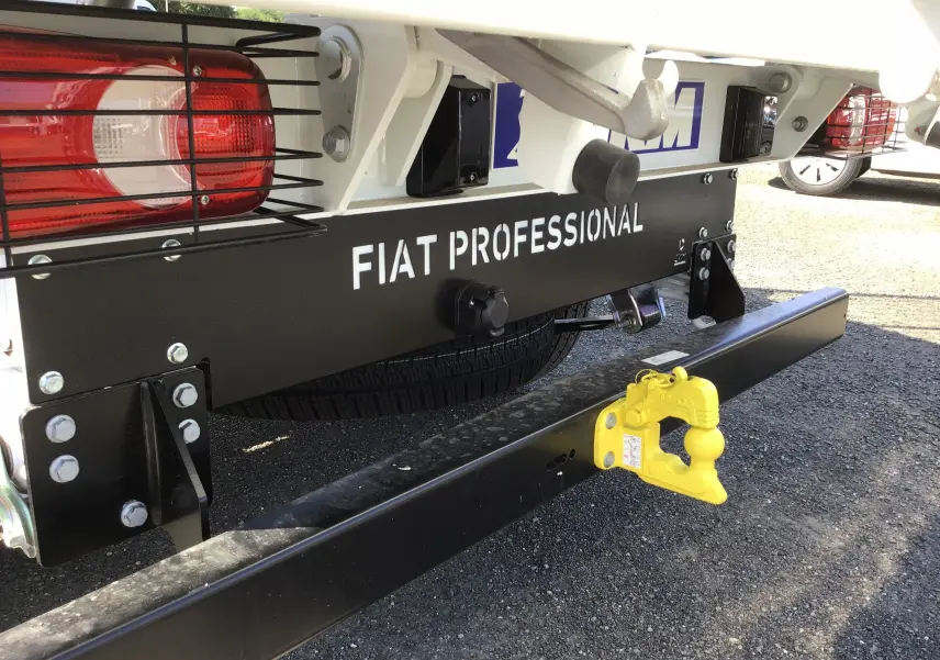 Gros plan arrière droit du FIAT Ducato blanc montrant la benne acier, le pare-chocs noir et l'attelage jaune vif.
